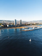 Vista aérea da orla de Barcelona, com a praia da Barceloneta, as torres do Port Olímpico e um veleiro no mar azul