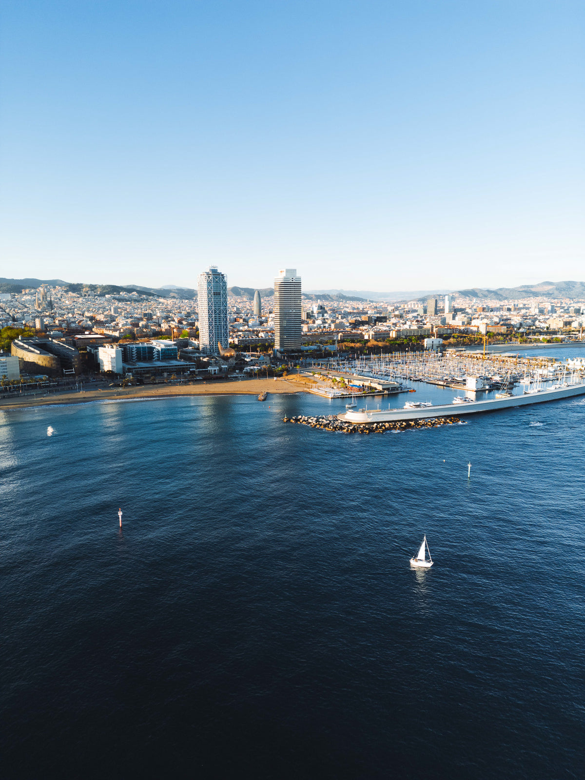 Vista aérea da orla de Barcelona, com a praia da Barceloneta, as torres do Port Olímpico e um veleiro no mar azul